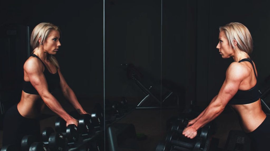 Athletic woman engaging in strength training exercise with dumbbells in a gym setting, reflecting on progress.